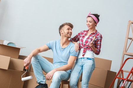 Young couple moving to new place man sitting on box holding paint roller looking at woman syanding with brush laughing cheerfulの写真素材