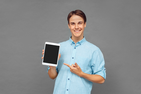 Freestyle. Teenager boy standing isolated on grey pointing at screen of digital tablet cheerfulの写真素材