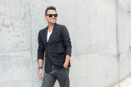 Outdoors leisure. Young man in suit and sunglasses on city street isolated on wall hand in pocket looking aside cheerfulの写真素材