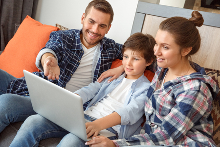 Family at home sitting on sofa in living room together searching info in internet on laptop pointing at screen joyfulの写真素材