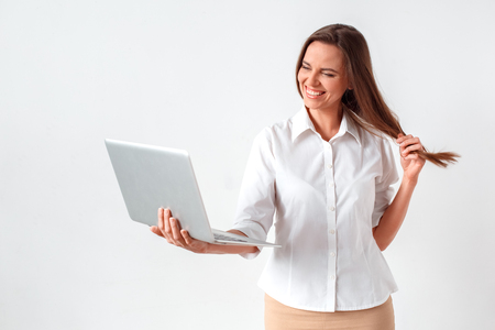 Online communication. Woman standing isolated on white video chatting on laptop touching hair laughing shyの写真素材