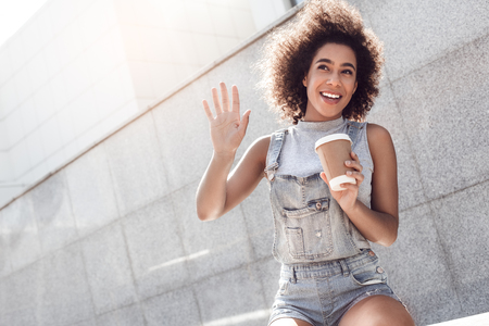 Young woman wearing overall shorts in the city street sitting drinking coffee waving to friend welcomingの写真素材