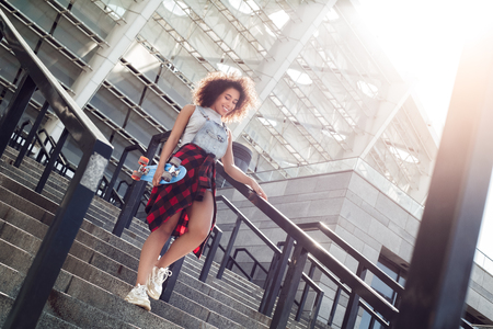 Young woman in the city street walking down the stairs with penny board looking down cheerful sunnyの写真素材
