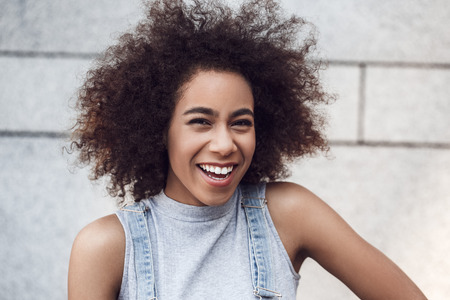 Young woman in the city street standing isolated on gray wall looking camera laughing cheerful close-upの写真素材