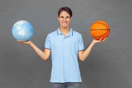 Freestyle. Teenager boy standing isolated on grey with globeand ball looking camera cheerfulの写真素材