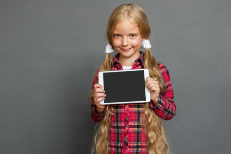 Little girl with ponytails standing isolated on grey showing screen of digital tablet looking camera playfulの写真素材