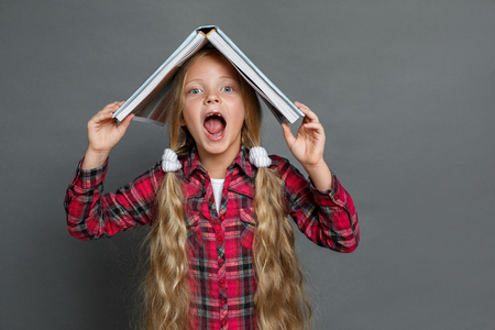 Little girl with ponytails standing isolated on grey with book on head shoutingの写真素材