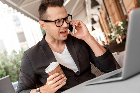 Lunch time. Young man in suit and glasses sitting at cafe on city street with laptop with cup of hot coffee shouting on smartphone irritated close-upの写真素材