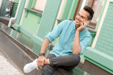 Outdoors leisure. Young man sitting on city street talking on smartphone smiling relaxedの写真素材