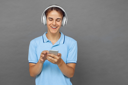 Music lover. Teenager boy in headphons standing isolated on grey choosing song on smartphone smiling excitedの写真素材