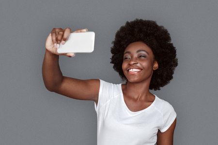 Young woman standing isolated on gray taking selfie on smartphone looking camera laughing cheerfulの写真素材