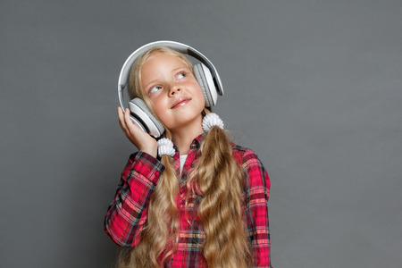 Little girl in headphones with ponytails standing isolated on grey listening song looking aside dreamfulの写真素材
