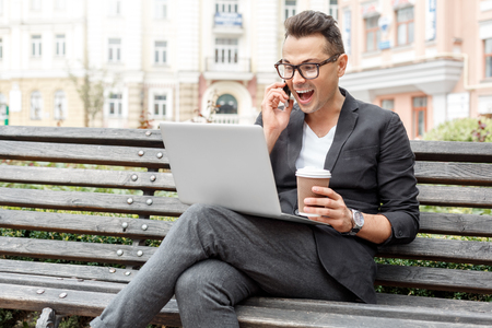 Outdoors leisure. Young man in suit and glasses on city street sitting on bench with laptop and cup of coffee talking on smartphone surprisedの写真素材