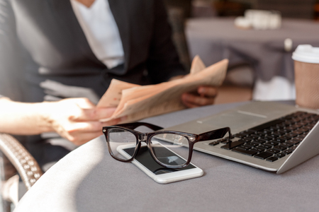 Lunch time. Young man in suit sitting at cafe on city street laptop smartphone and eyeglasses close-up blurred backgroundの写真素材