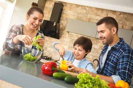 Family at home standing in kitchen together parents laughing looking at boy cutting vegetables for saladの写真素材