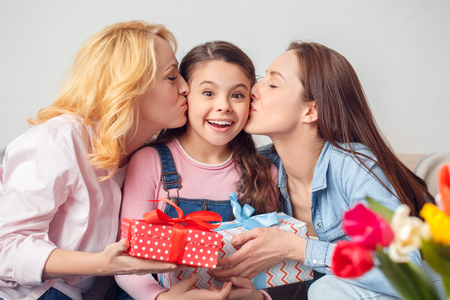 Grandmother mother and daughter together at home celebration sitting women kissing girl with present surprisedの写真素材