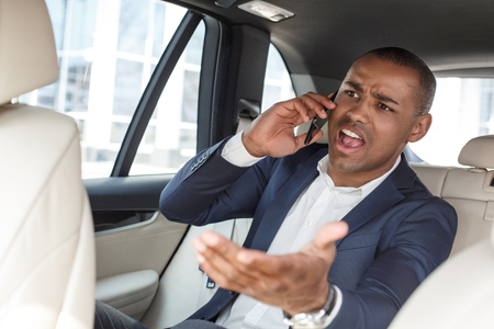 Young businessman sitting on back seat in car talking on smartphone shouting at driver irritatedの写真素材