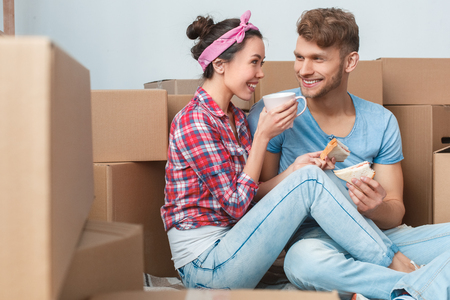 Young couple moving to new place sitting drinking tea eating sandwiches talking smiling joyful side viewの写真素材