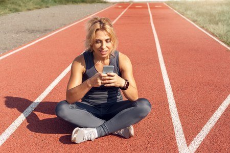 Sporty Lifestyle. Young woman on stadium sitting on track checking social media on smartphone smiling happyの写真素材