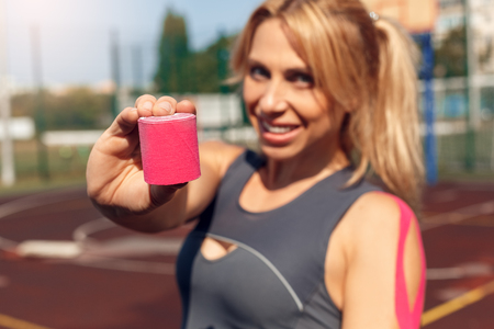 Sporty Lifestyle. Young woman with kinesio tape standing on stadium with tape roll close-up blurred smiling happyの写真素材