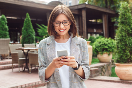 Business casual. Young lady in eyeglasses standing on the city street using app on smartphone happyの写真素材