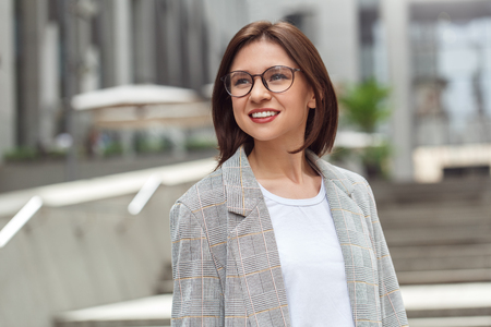 Business casual. Young lady in eyeglasses standing on the city street smiling cheerful close-upの写真素材