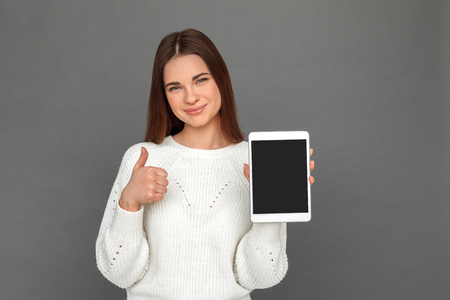Freestyle. Young girl standing isolated on grey showing screen of digital tablet thumb up smiling happyの写真素材