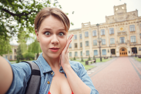 Young student with backpack at university campus standing taking selfie on smartphone looking camera surprised close-upの写真素材