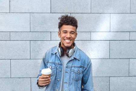 Freestyle. Mulatto guy with headphones on neck standing isolated on wall with cup of coffee smiling toothyの写真素材