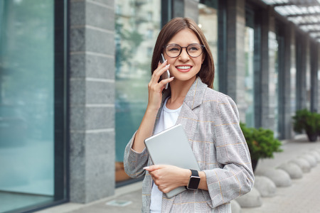 Business communications. Young lady in eyeglasses standing on the city street with digital tablet talking on smartphone cheerfulの写真素材