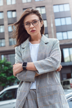 Business casual. Young lady in eyeglasses standing on the city street crossed arms looking camera seriousの写真素材
