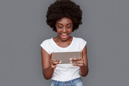 Young woman standing isolated on gray with digital tablet watching video shouting excitedの写真素材