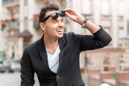 Outdoors leisure. Young man in suit standing on city street taking sunglasses off smiling surprised close-upの写真素材
