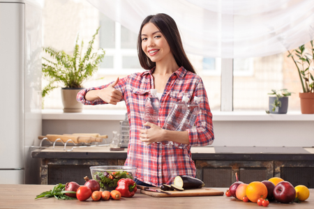Young girl at kitchen healthy lifestyle standing with water bottles looking camera cheerful showing thumb upの写真素材