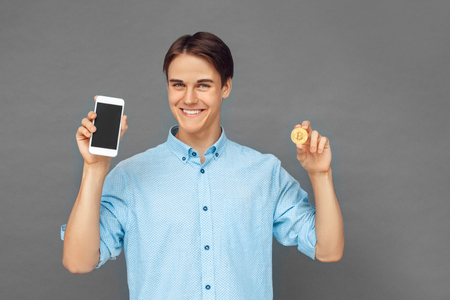 Freestyle. Teenager boy standing isolated on grey with cryptocurrency coin and smartphone looking camera joyfulの写真素材