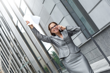 Business woman in eyeglasses standing on the city street answering call hand up with contract cheerfulの写真素材