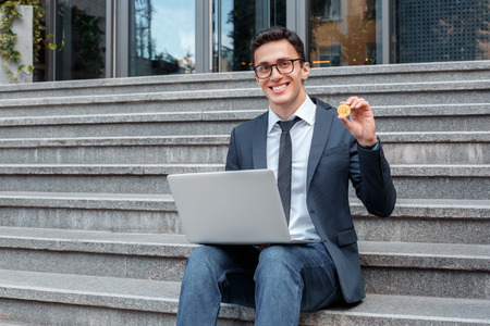 Electronic cash. Businessman in eyeglasses sitting on stairs on the city street working on laptop showing coin smiling happyの写真素材