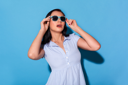 Freestyle. Woman in dress wearing eyeglasses standing isolated on blue wall smiling glamorousの写真素材