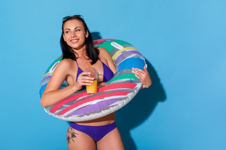 Beach style. Woman in bikini wearing swim ring and sunglasses up the head standing isolated on blue wall drinking smoothie laughing playfulの写真素材