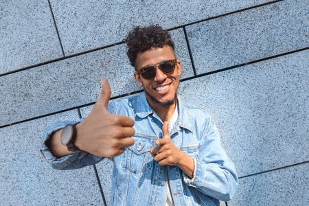 Freestyle. Mulatto guy in sunglasses standing isolated on wall showing thumb up smiling cheerful close-upの写真素材