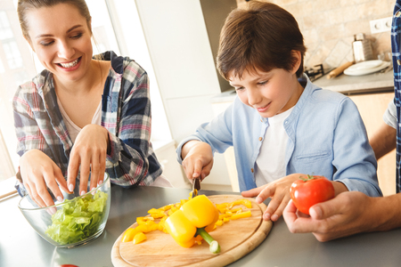 Family at home standing in kitchen making salad together son cutting vegetables concentrated smiling excited close-upの写真素材