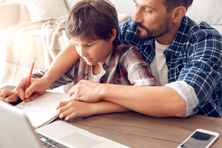 Father and son at home sitting at table doing assignment dad holding boys hand writing solution close-upの写真素材