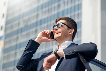 Young man in sunglasses leaning on car door talking on smartphone looking up dreamful bottom viewの写真素材