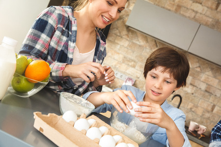 Family at home standing near table in kitchen together mother and son beaking eggs cheerful close-upの写真素材
