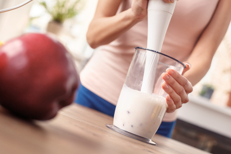 Young girl at kitchen healthy lifestyle standing blending smoothie with hand blender close-upの写真素材