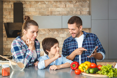 Family at home standing in kitchen together mother and son looking concentrated at father showing video on digital tablet cheerfulの写真素材