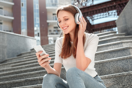 Young girl in headphones sitting on stairs in the city with smartphone listening music closed eyes joyfulの写真素材