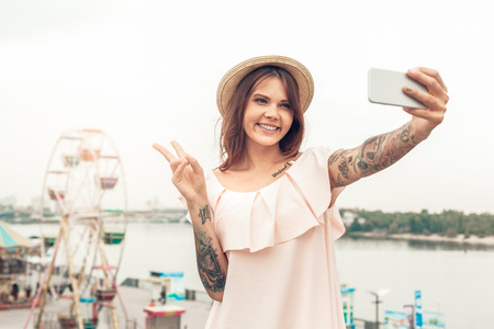 Ourdoors Leisure. Girl in hat standing near seafront taking selfie on smartphone showing peace...