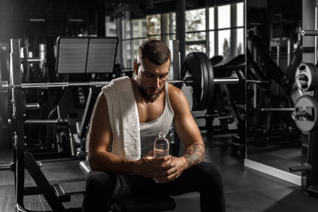Bodybuilding. Bearded man sitting on bench at gym with bottle of water looking down tiredの写真素材