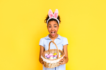 Easter Celebration Concept. Young woman in cute dress and bunny ears standing isolated on yellow with basket of eggs screaming excitedの写真素材